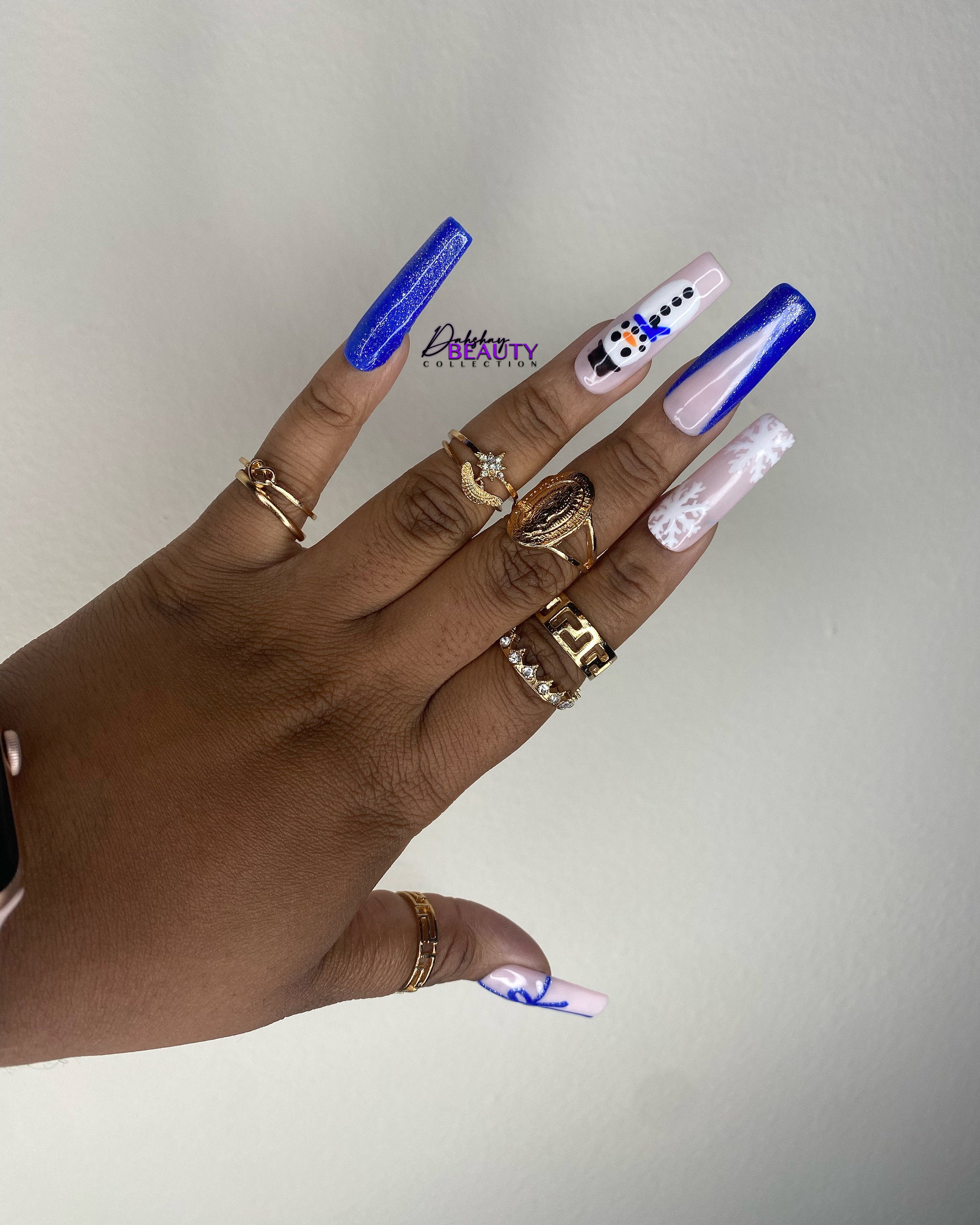 Hand with Christmas nails that has a snowman, snowflake and a bow. The hand also has multiple gold rings on a plain background.