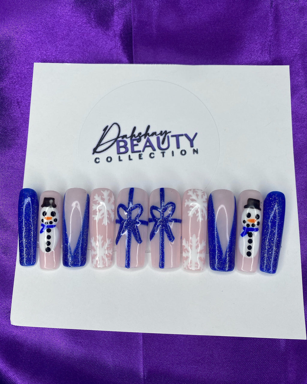 Press On Nails with Christmas designs of snowmen, bows  and a snowflake on a white card against a purple satin background.