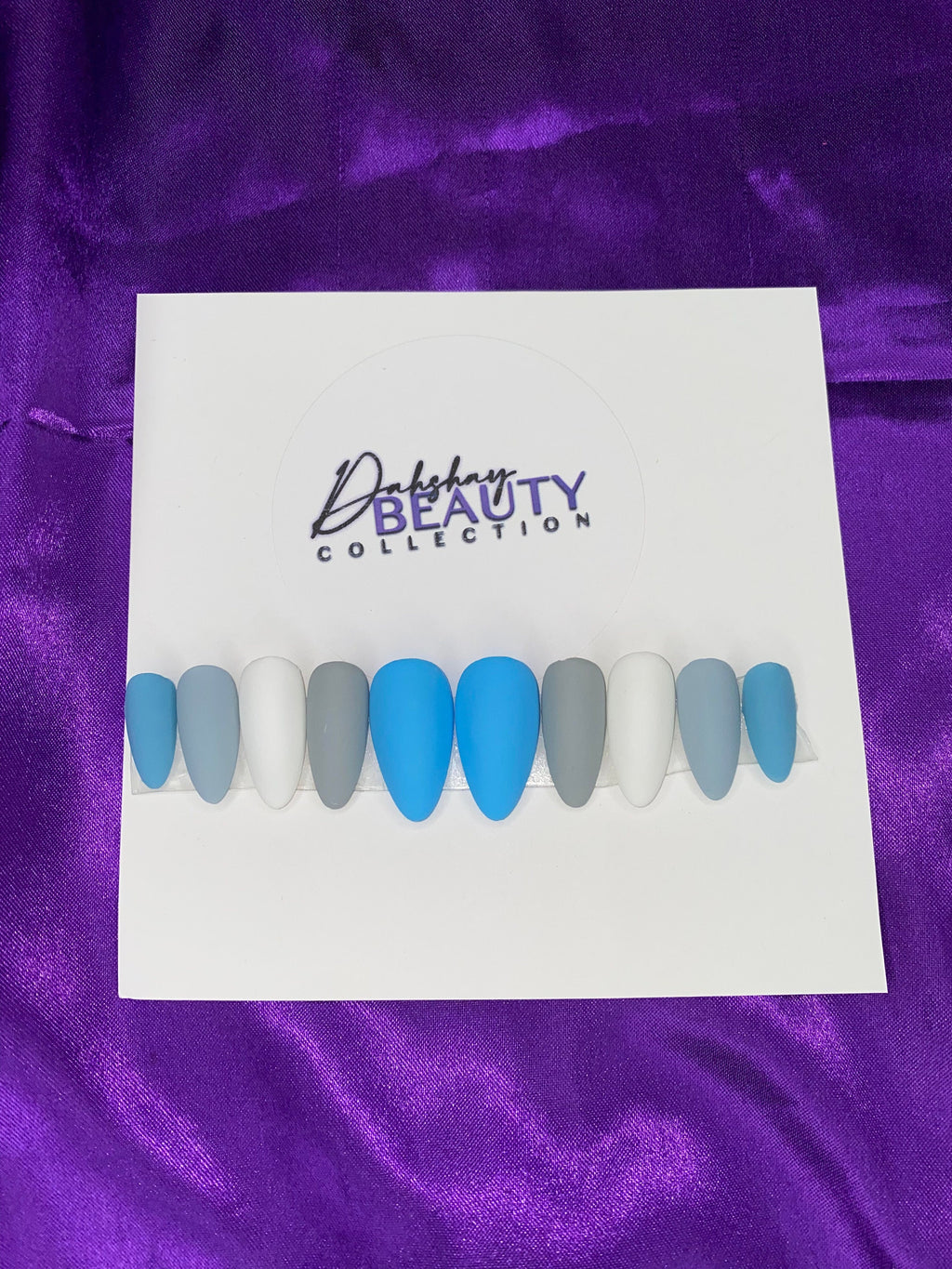 Set of blue, grey and white nails on while paper with a purple satin background.