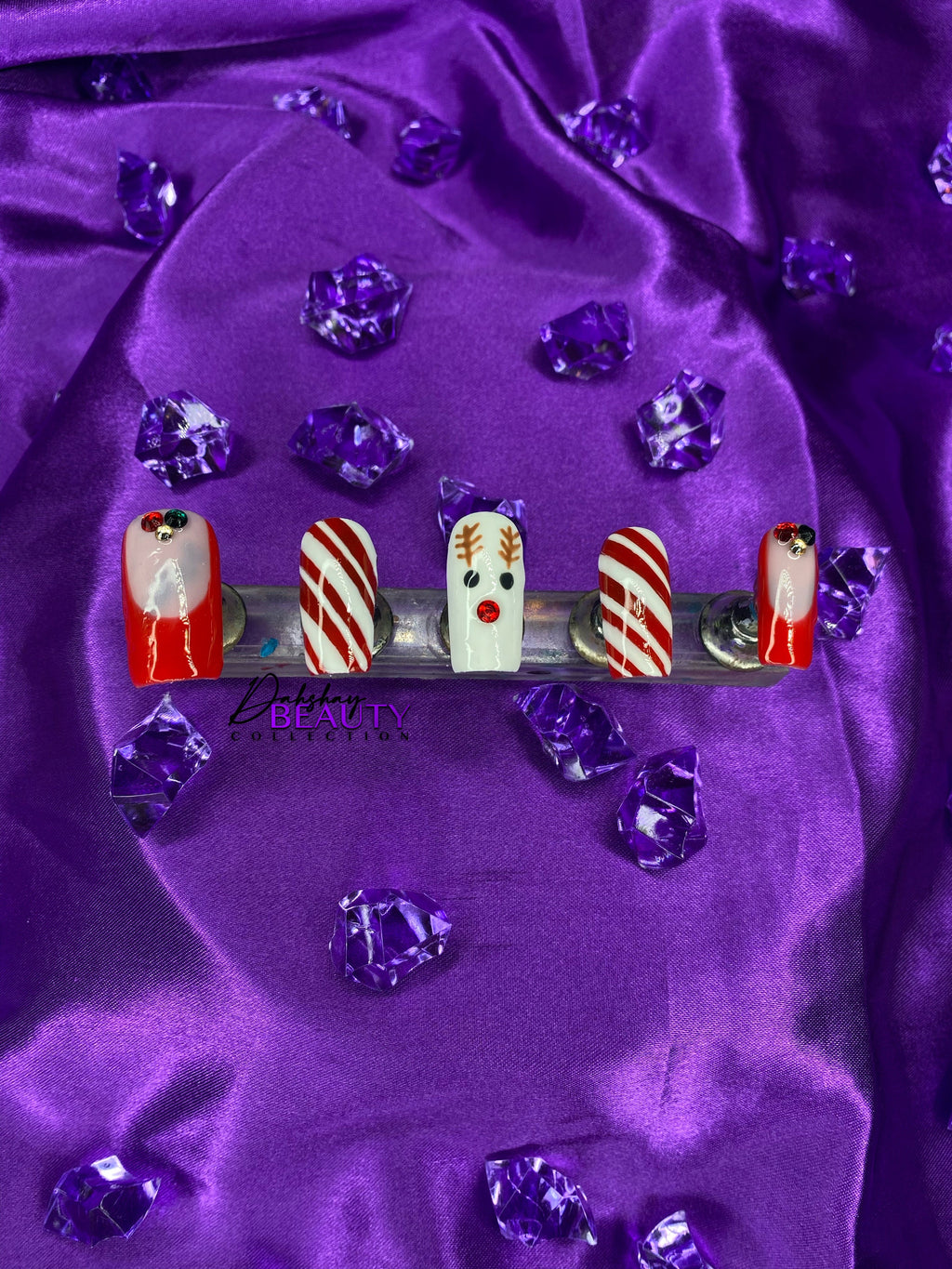 Nail art set with reindeer and candy cane designs on a purple fabric background with purple diamonds, featuring the Dahshay Beauty Collection logo.