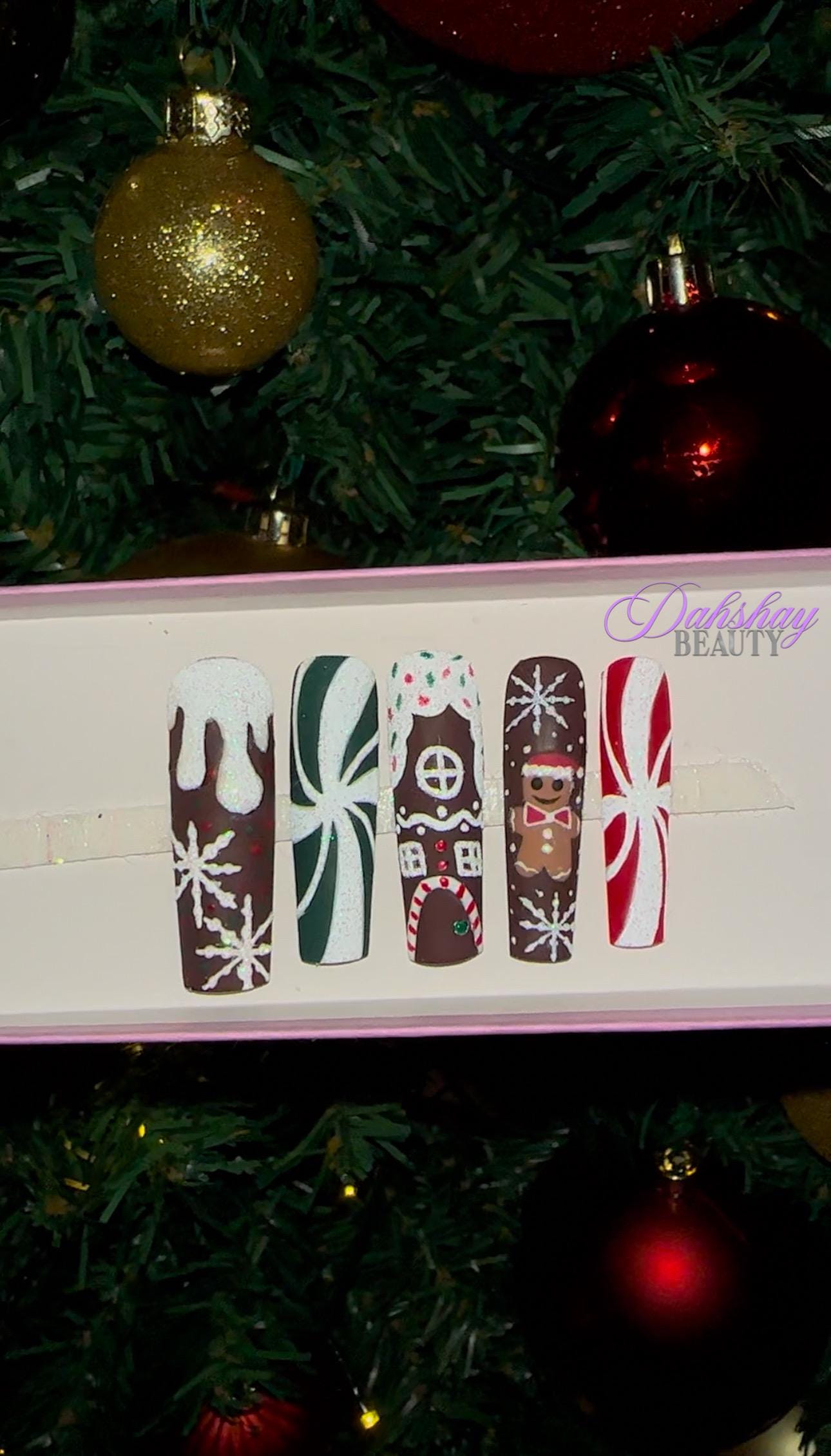 Set of Christmas gingerbread themed nail art designs on a white tray with a decorated Christmas tree in the background.