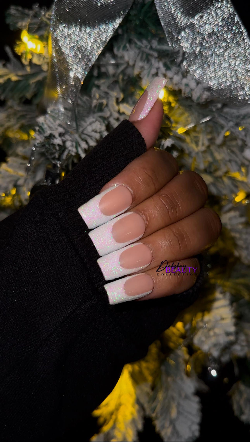Hand wearing a black long sleeve jacket with white glitter french tip nails against a Christmas tree background