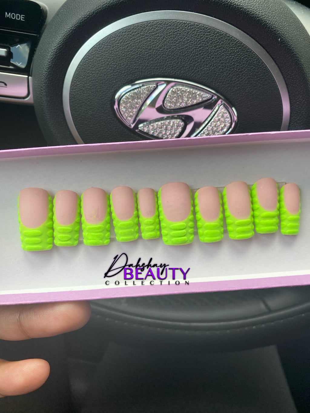 Display of false nails with green nails with 3D stripes held by a hand, against a car interior background.