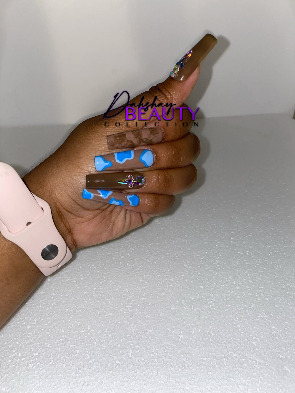 Blue and brown nails on a hand with a watch in front of a plain background.