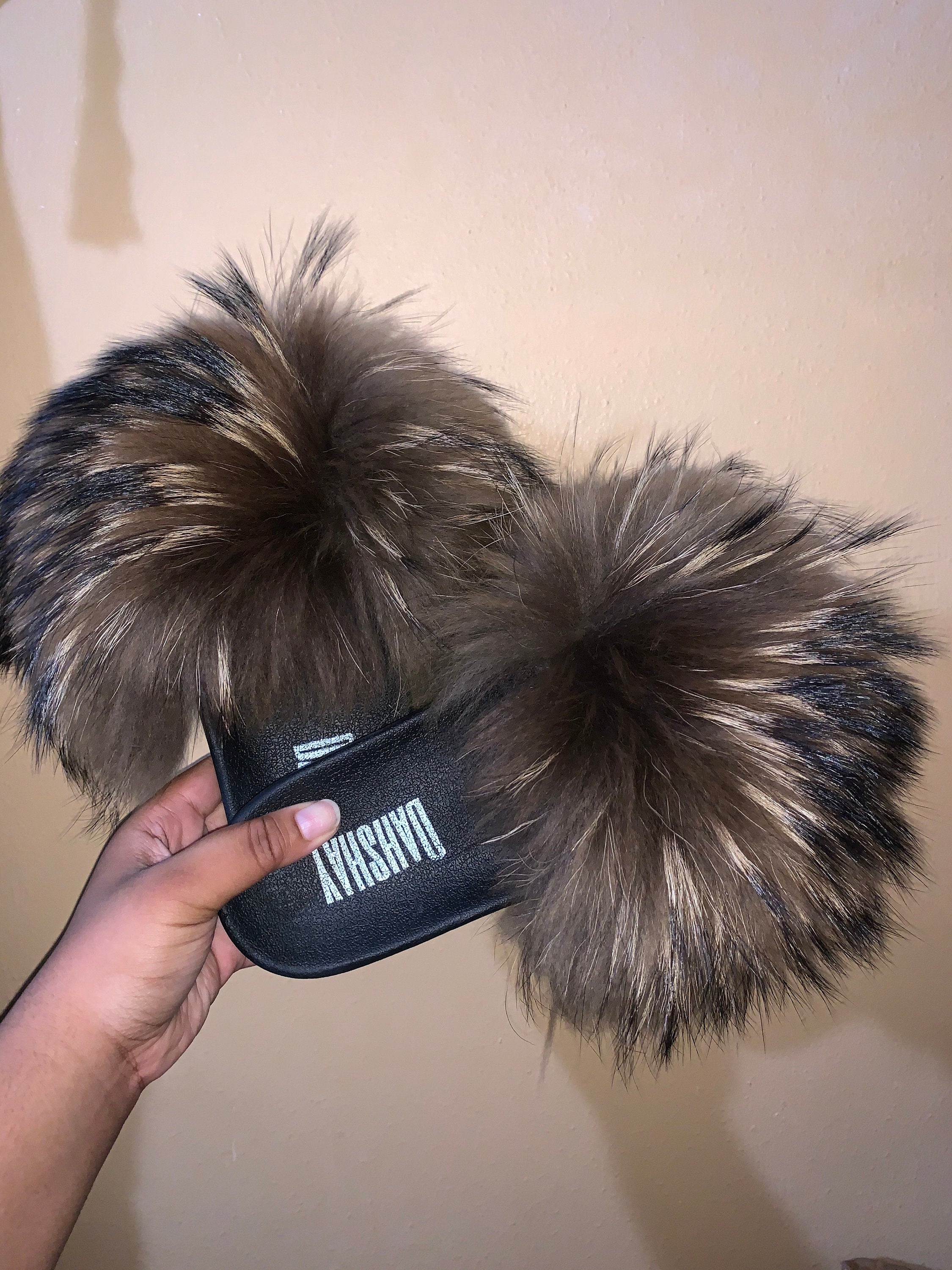 Fuzzy slippers with a brand logo held by a hand against a plain background