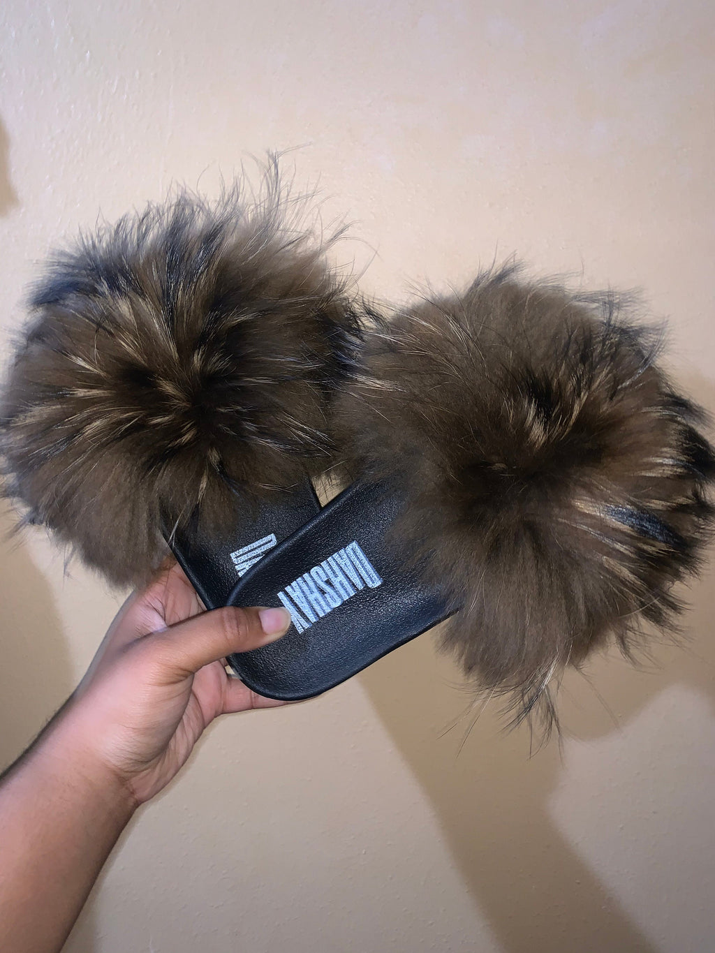 Fuzzy slippers with a brand logo held by a hand against a plain background