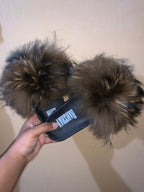 Fuzzy slippers with a brand logo held by a hand against a plain background