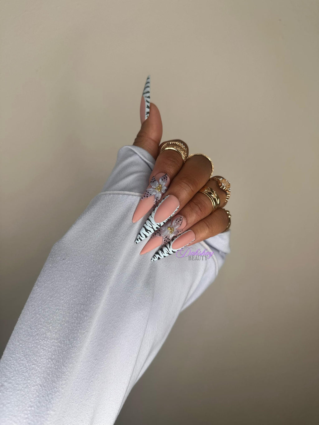 Hand with floral nail design wearing a light blue sleeve against a beige background with gold rings