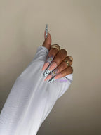 Hand with floral nail design wearing a light blue sleeve against a beige background with gold rings