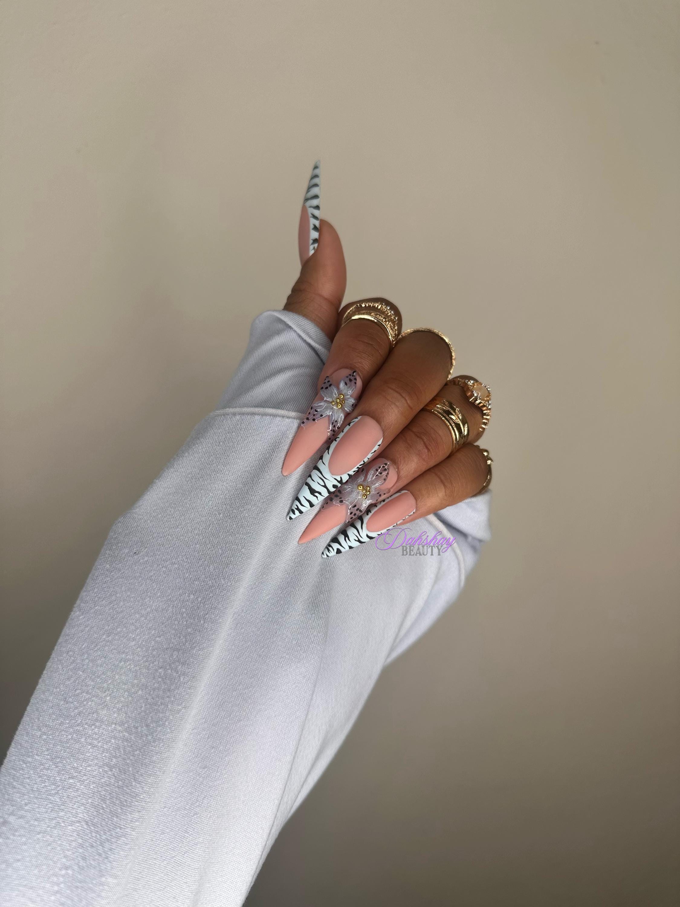 Hand with floral nail design wearing a light blue sleeve against a beige background with gold rings