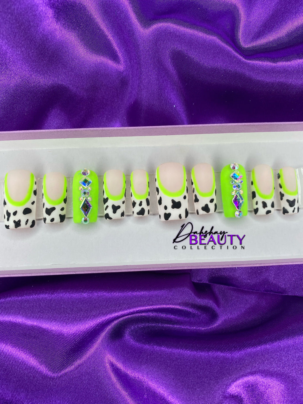 Set of decorative false nails with cow print and green accents on a purple fabric background