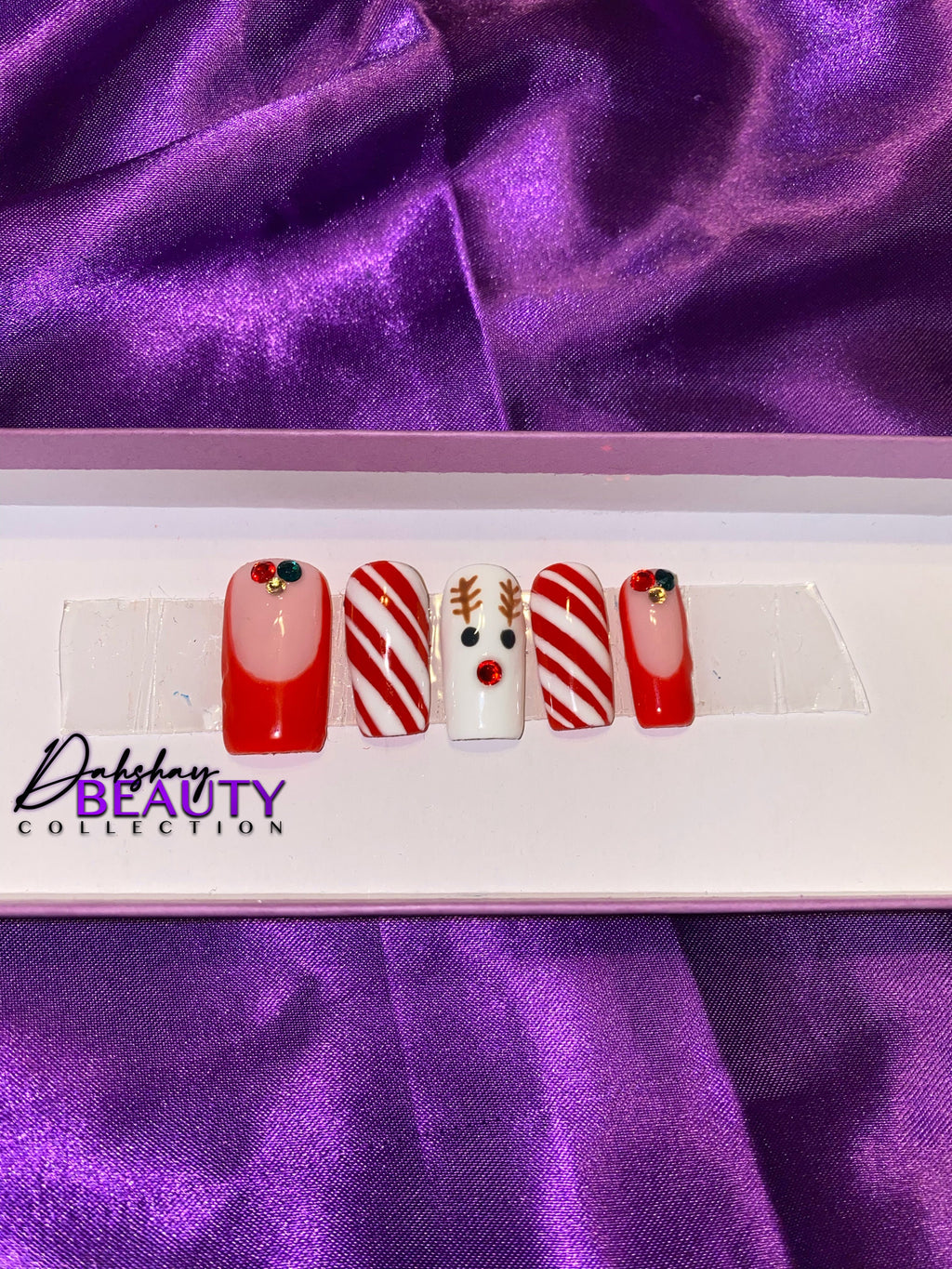 Nail art set with reindeer and candy cane designs on a purple fabric background, featuring the Dahshay Beauty Collection logo.