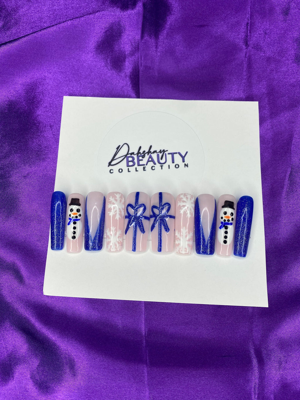 Press On Nails with Christmas designs of snowmen, bows  and a snowflake on a white card against a purple satin background.