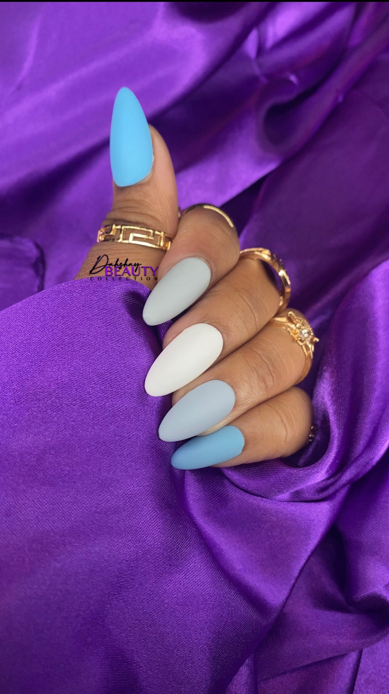 Close-up of a hand with blue, white and gray nails against a purple fabric background with gold rings on hand.