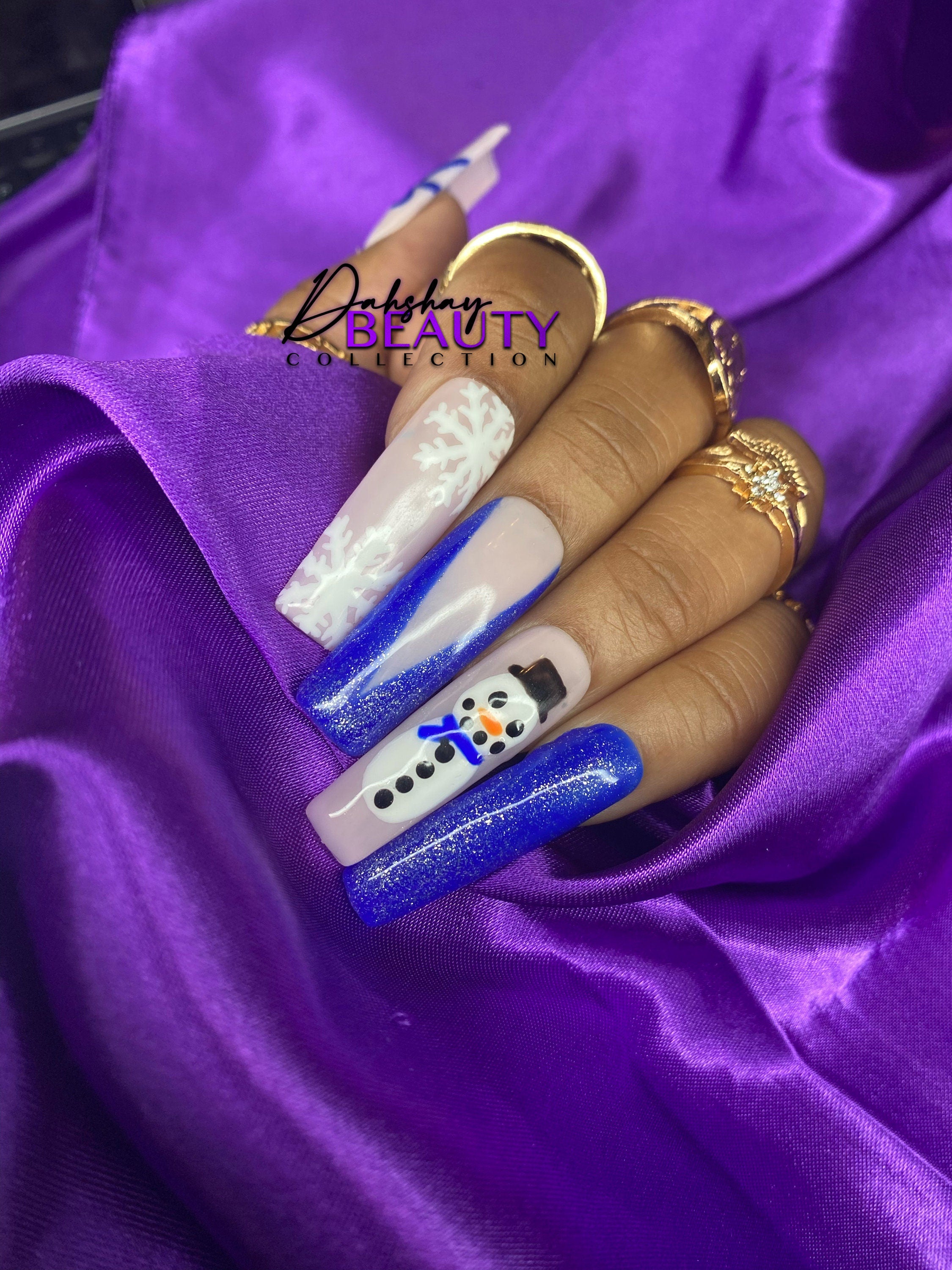 Press On Nails with Christmas designs of snowmen, bows  and a snowflake on a hand against a purple satin background.