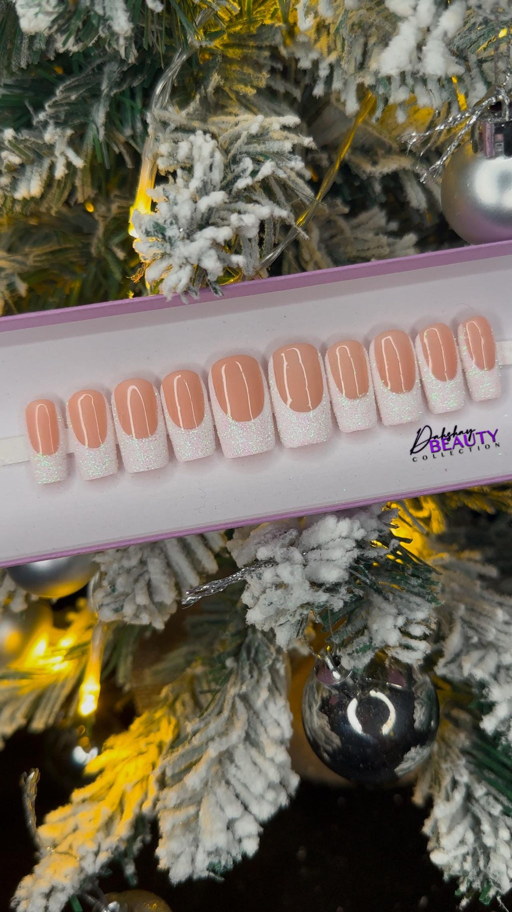 A set of nude false nails with a glitter white french tip on a display stand against a Christmas tree background