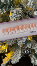 A set of nude false nails with a glitter white french tip on a display stand against a Christmas tree background