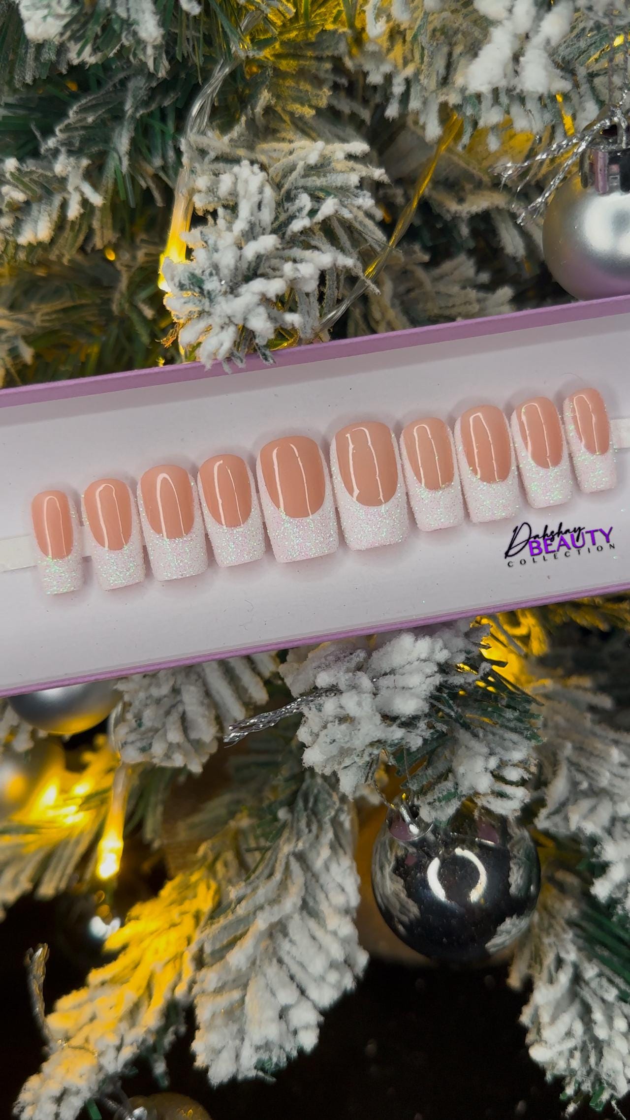 A set of nude false nails with a glitter white french tip on a display stand against a Christmas tree background