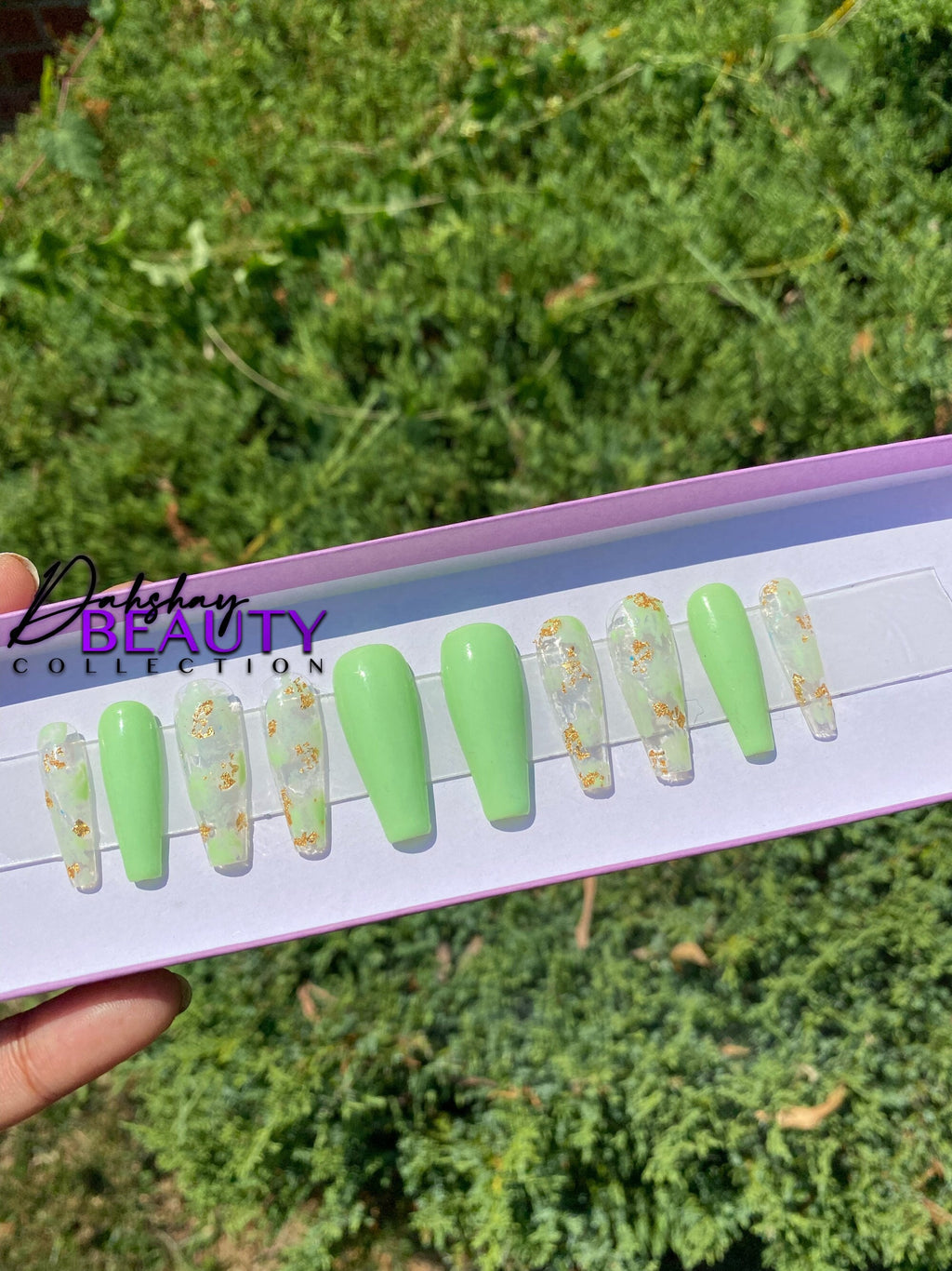 Set of green press on nails with gold designs on a display card against a green outdoor background.