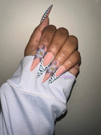 Hand with floral nail design wearing a light blue sleeve against a beige background with gold rings