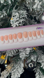 A set of nude false nails with a glitter white french tip on a display stand against a Christmas tree background