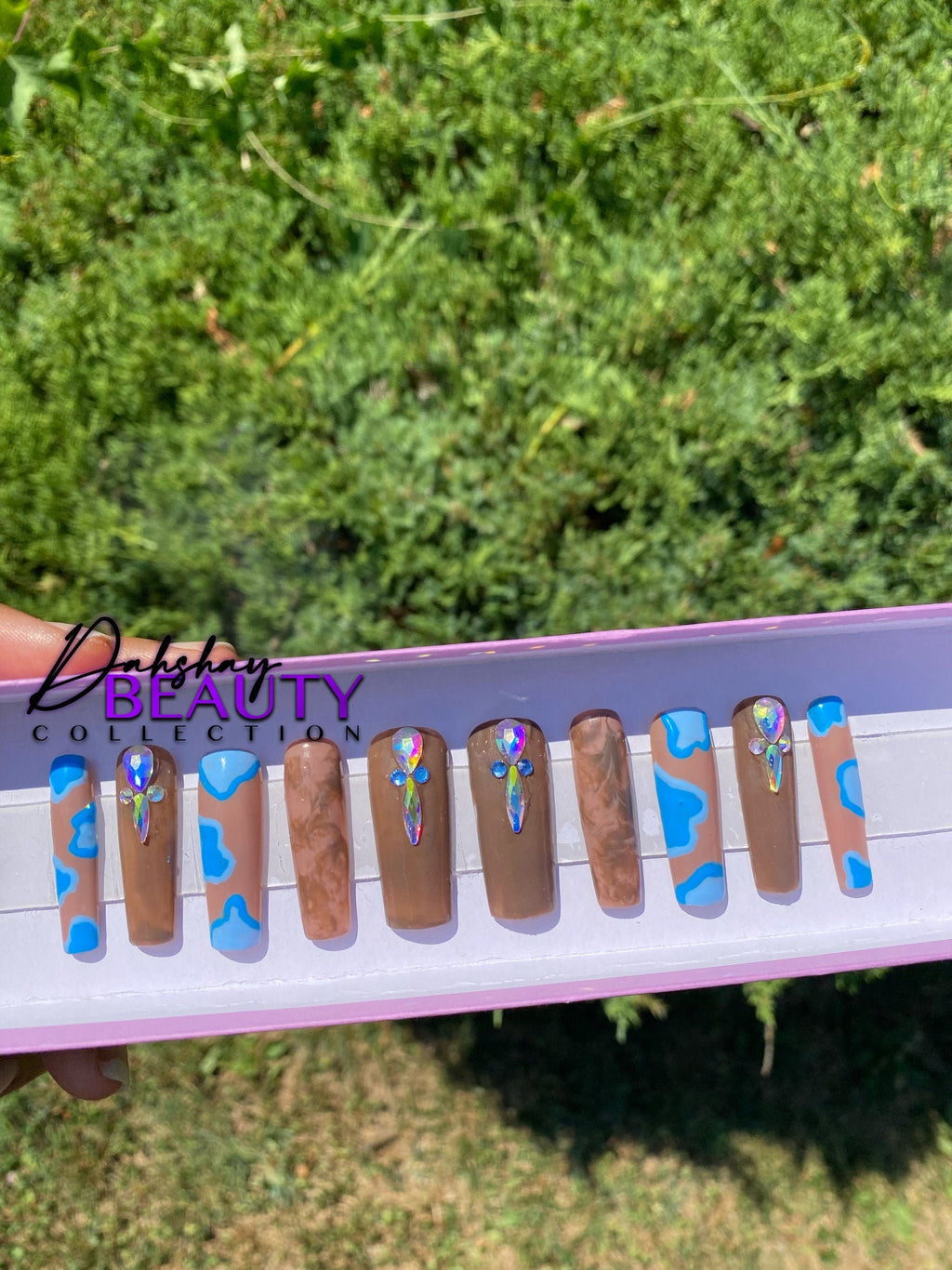 Brown nails with a blue abstract design with rhinestone in a tray help against a green out door tree