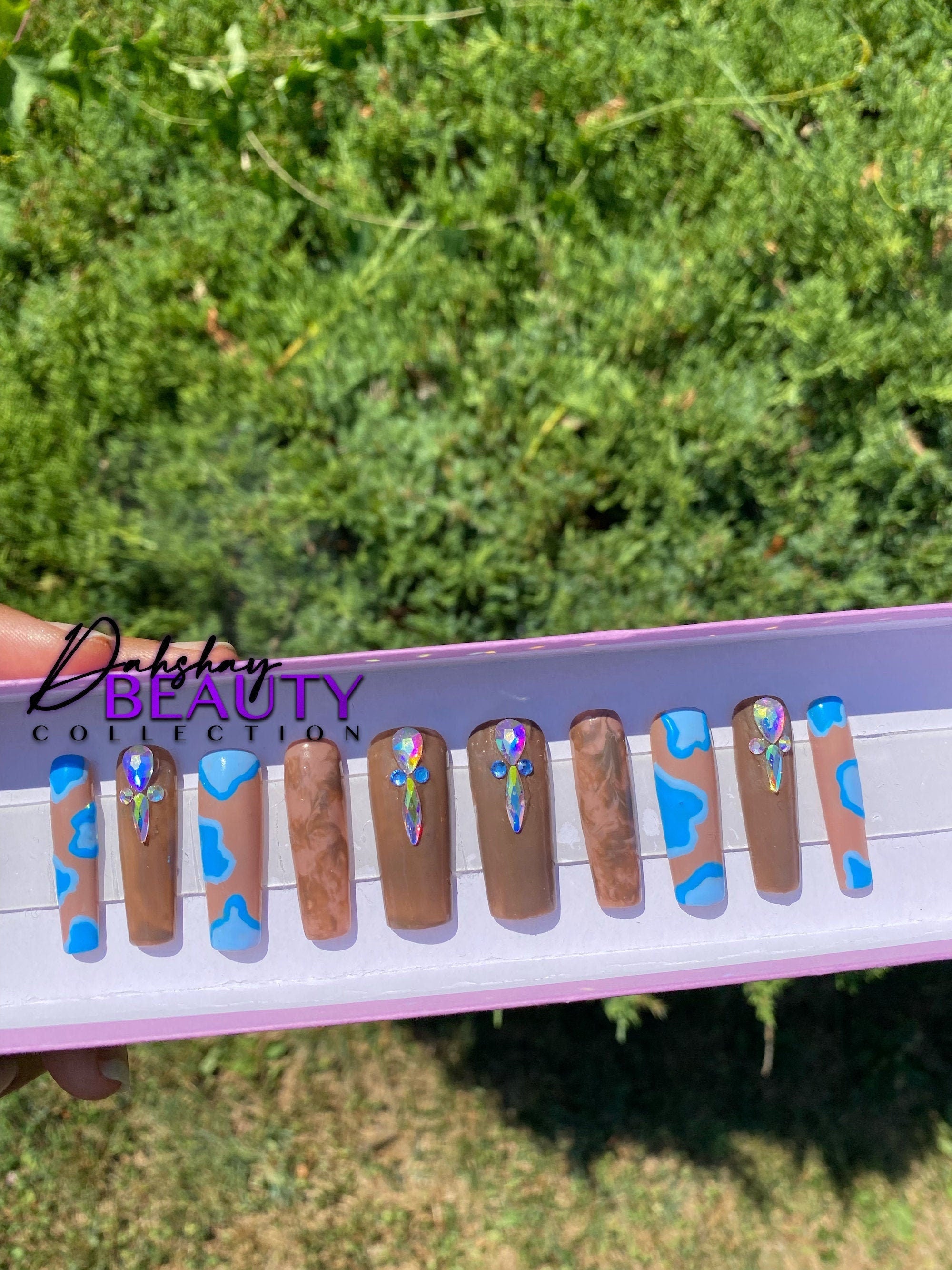 Brown nails with a blue abstract design with rhinestone in a tray help against a green out door tree