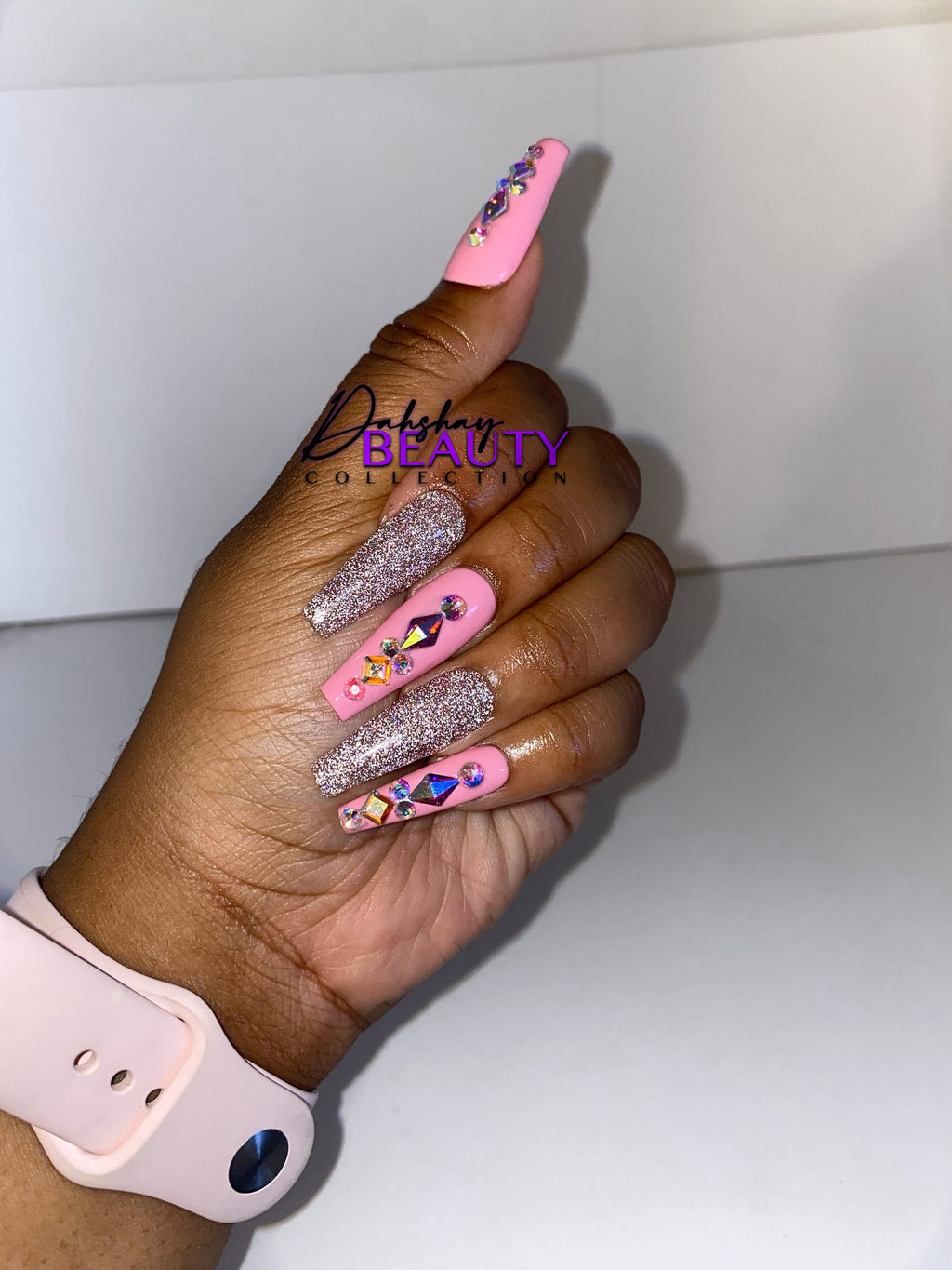 Hand with pink, rhinestones and glittery nails, wearing a light pink smartwatch, on a white background.