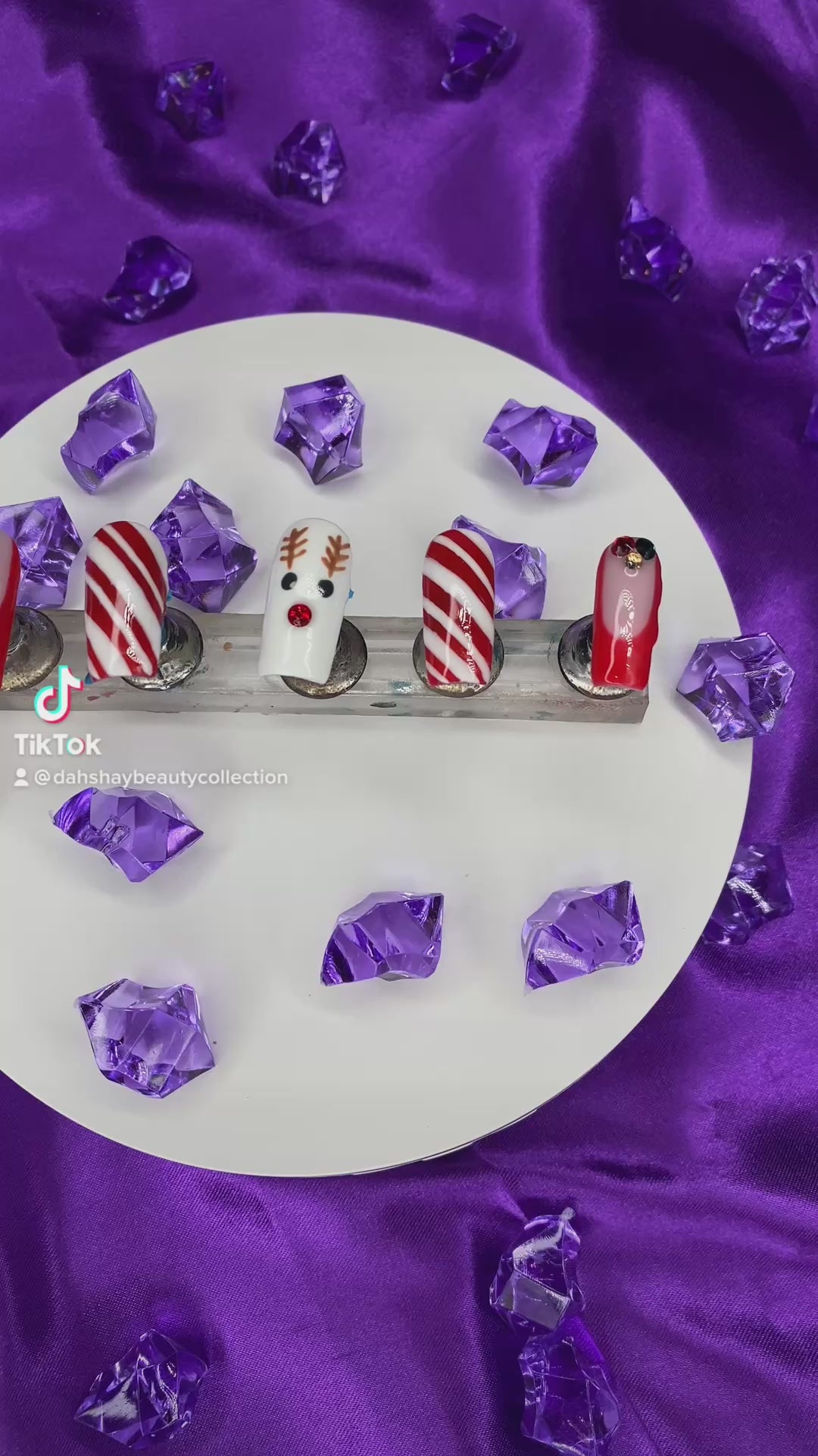 A video of a set of Christmas nails with a reindeer, candy cane and french tip design on a purple satin background with purple diamonds.
