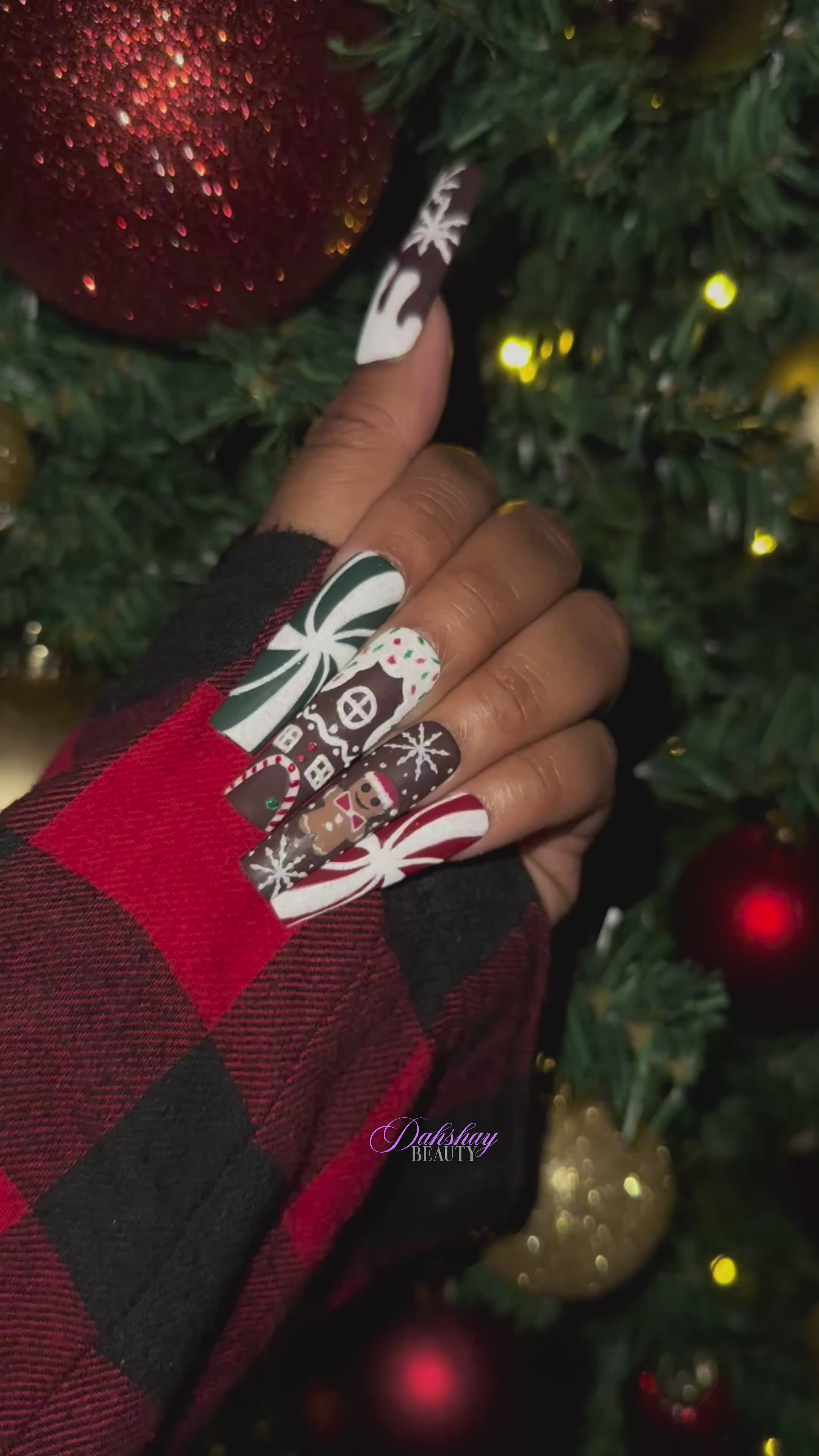 A video of a set of Christmas gingerbread themed nail art designs on a hand with a decorated Christmas tree in the background.