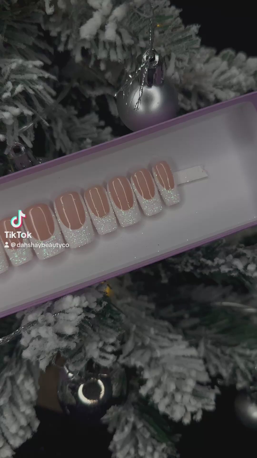 A video of nude false nails with a glitter white french tip on a display stand against a Christmas tree background