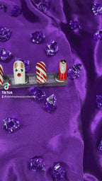 A video of a set of Christmas nails with a reindeer, candy cane and french tip design on a purple satin background with purple diamonds.