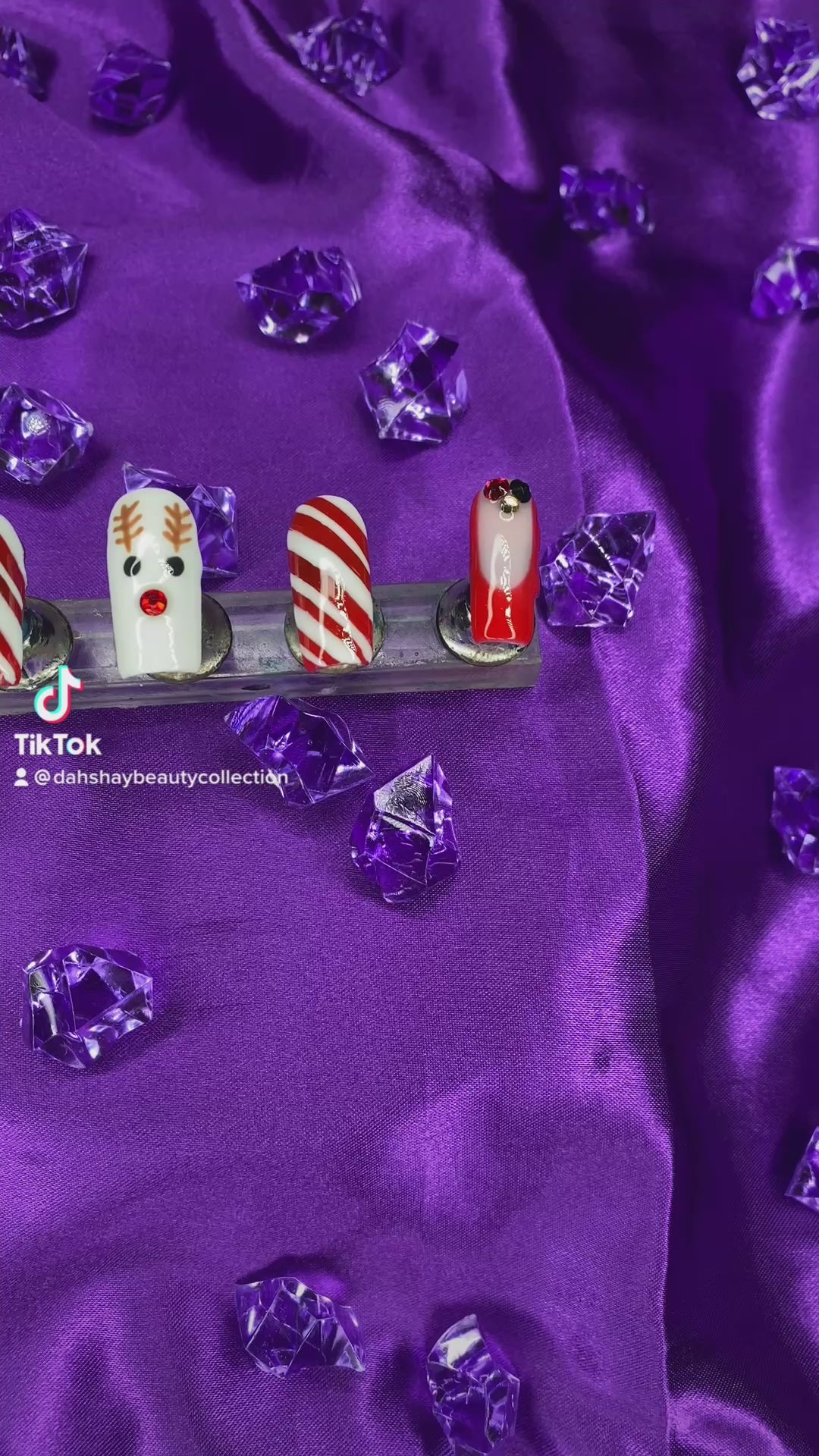 A video of a set of Christmas nails with a reindeer, candy cane and french tip design on a purple satin background with purple diamonds.