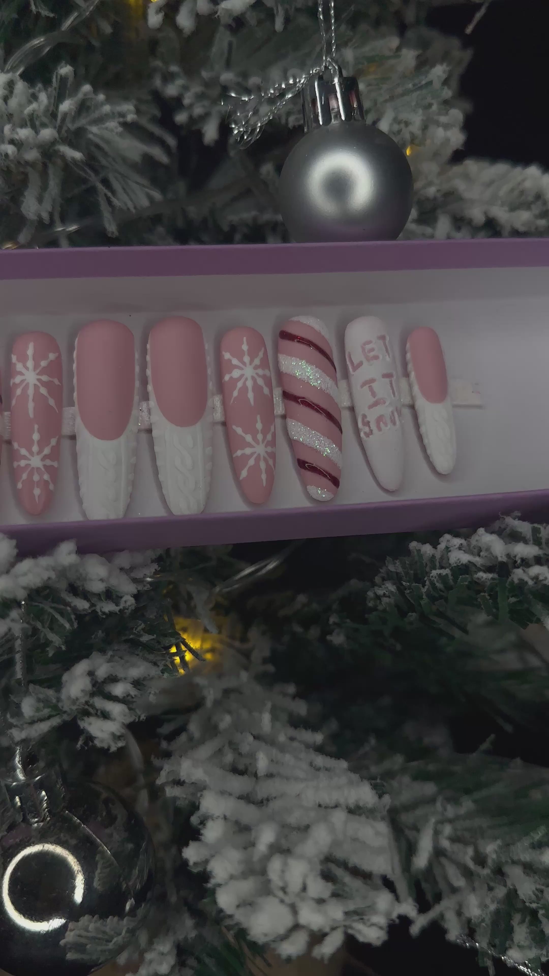 A video of a set of Christmas false nails with snowflakes, candy cane and sweater designs in front of a Christmas tree.
