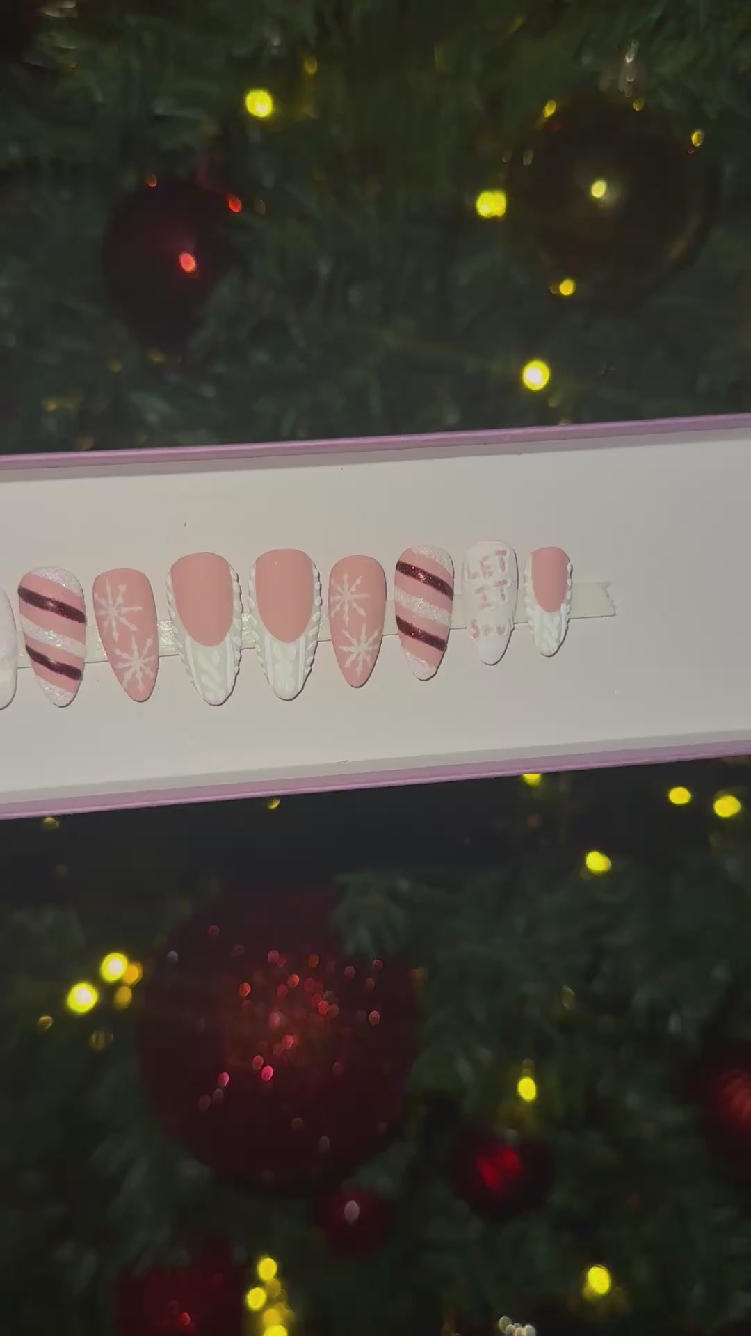 A video of a set of Christmas false nails with snowflakes, candy cane and sweater designs in front of a Christmas tree with red and gold ornaments