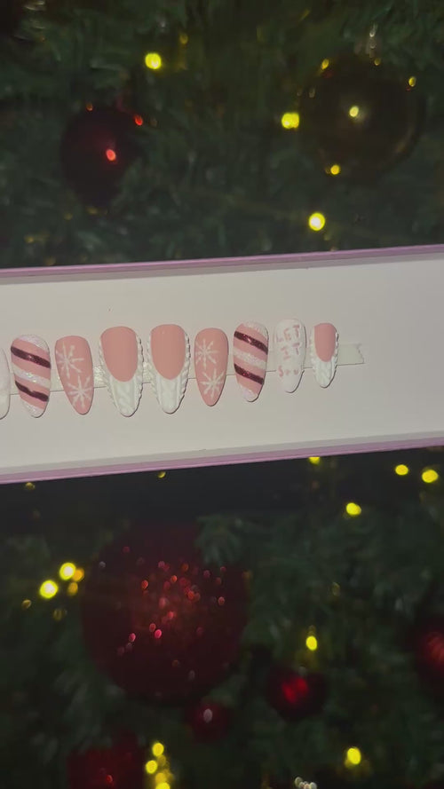 A video of a set of Christmas false nails with snowflakes, candy cane and sweater designs in front of a Christmas tree with red and gold ornaments