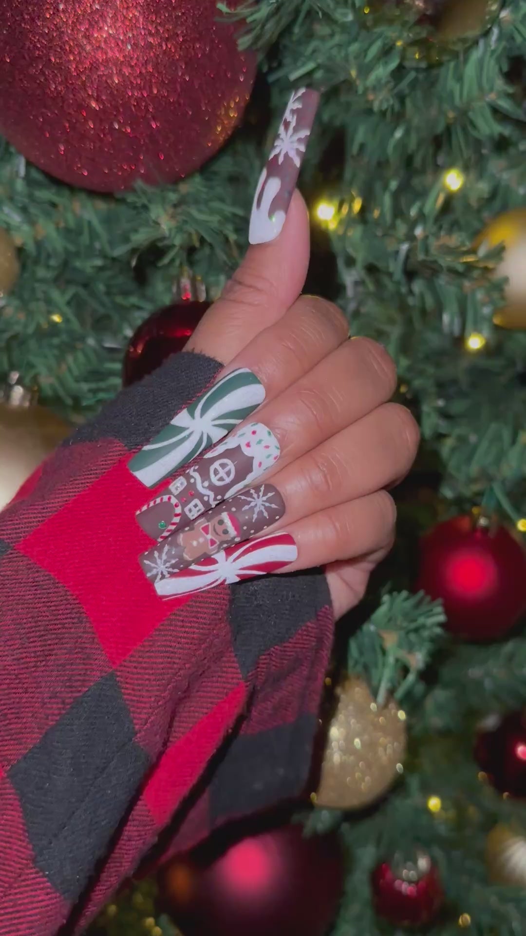 A video of a set of Christmas gingerbread themed nail art designs on a hand with a decorated Christmas tree in the background.