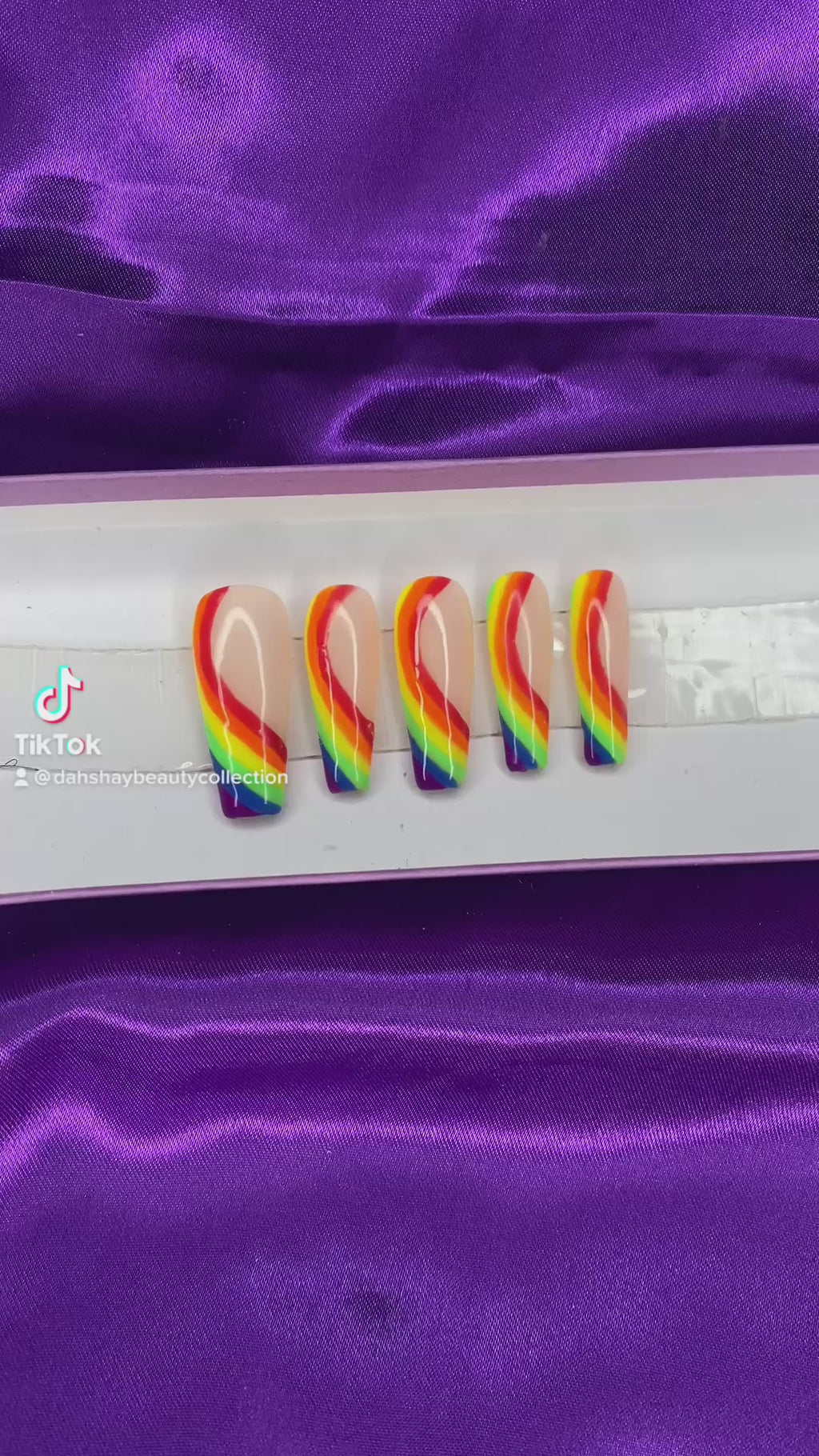 A video of rainbow-colored press-on nails on a white tray against a purple satin background.