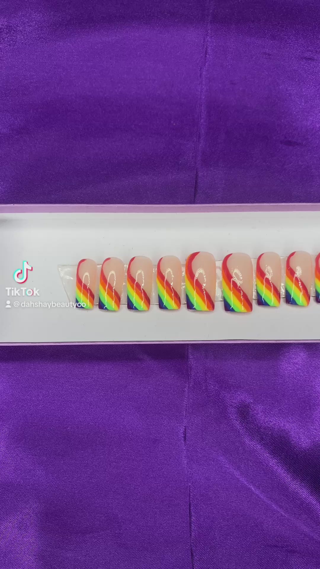 A video of rainbow-colored press-on nails on a white tray against a purple satin background.