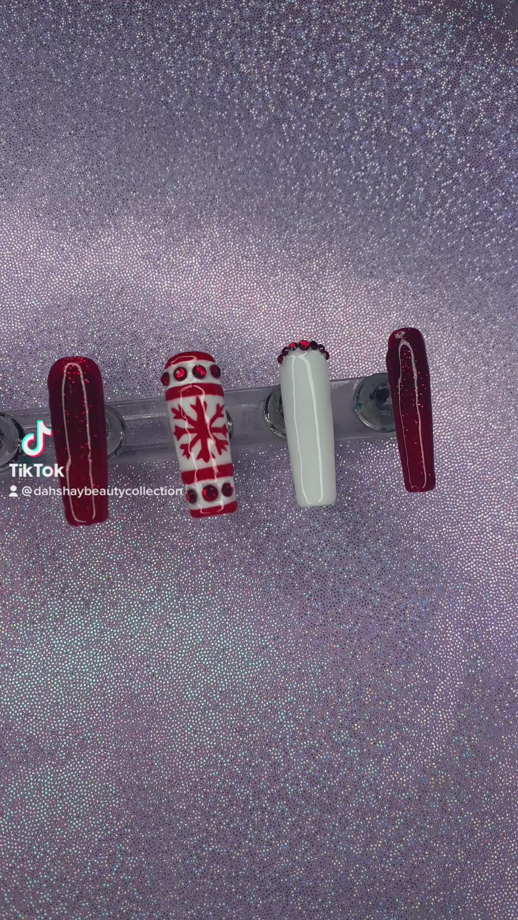 A video of a set of red and white Christmas nail extensions with red glitter and a snowflake design on a purple glittery background.