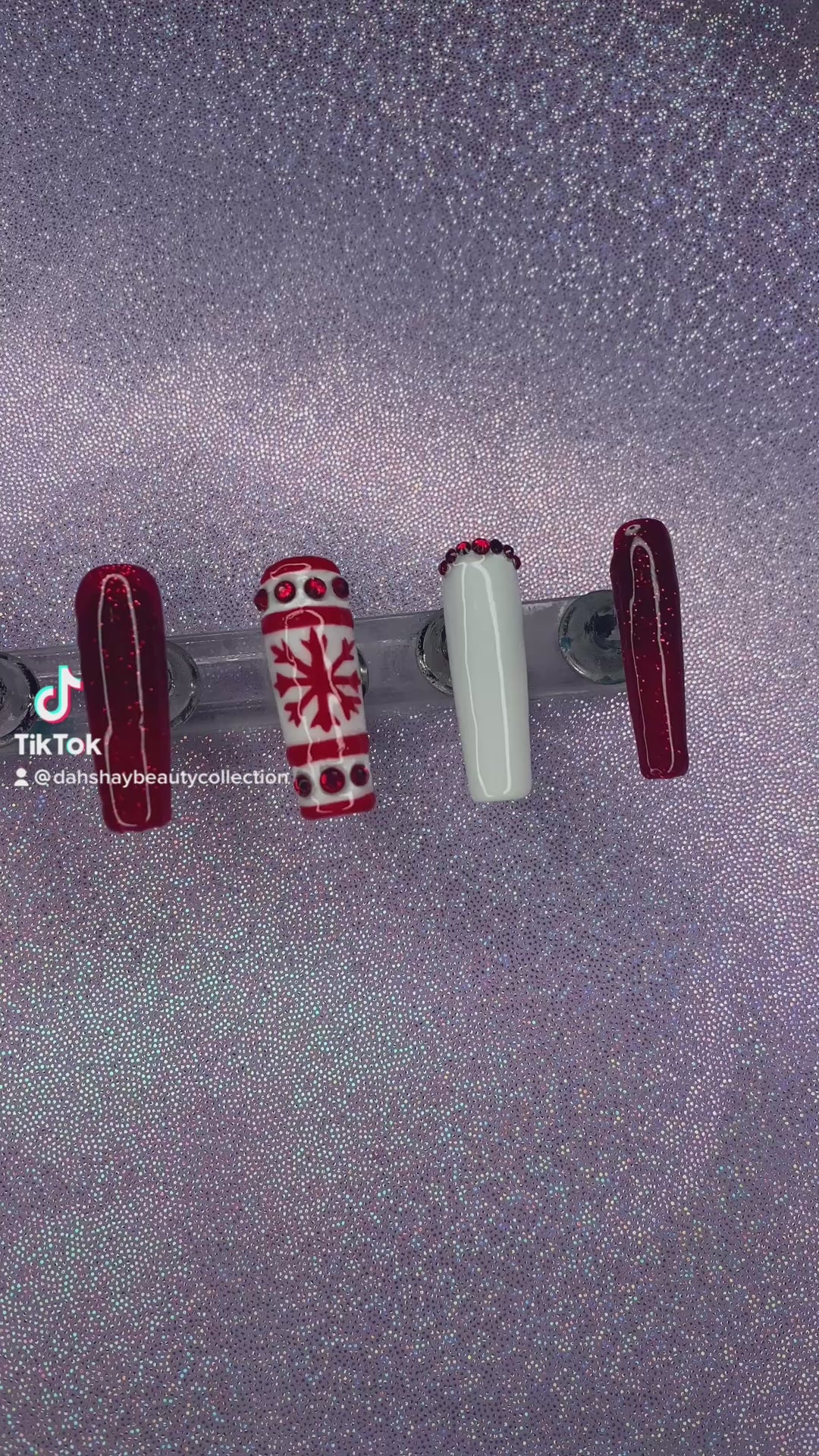 A video of a set of red and white Christmas nail extensions with red glitter and a snowflake design on a purple glittery background.